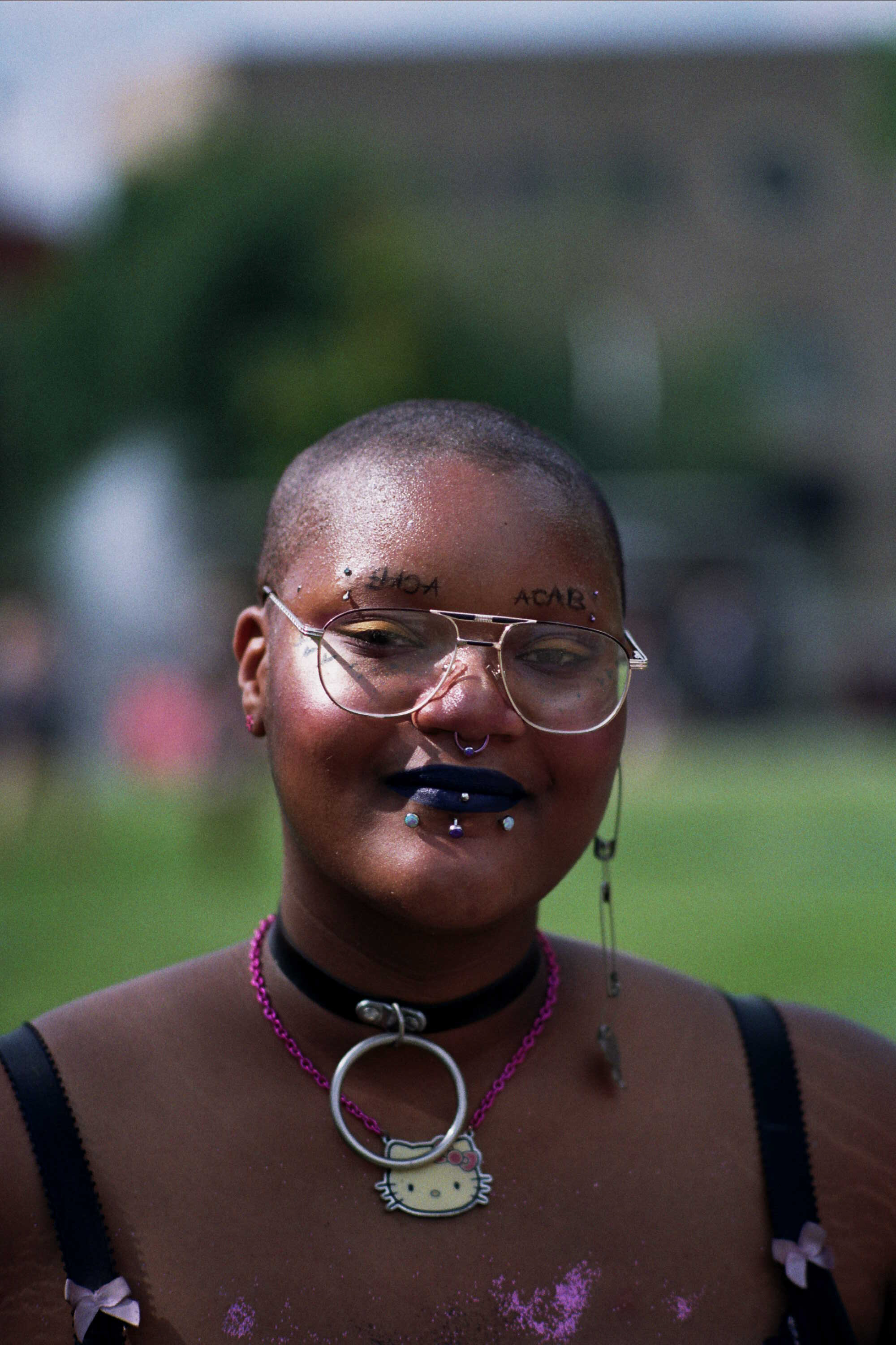 A person with a shaved head, dark lipstick, and a Hello Kitty necklace smiles gently at the camera.