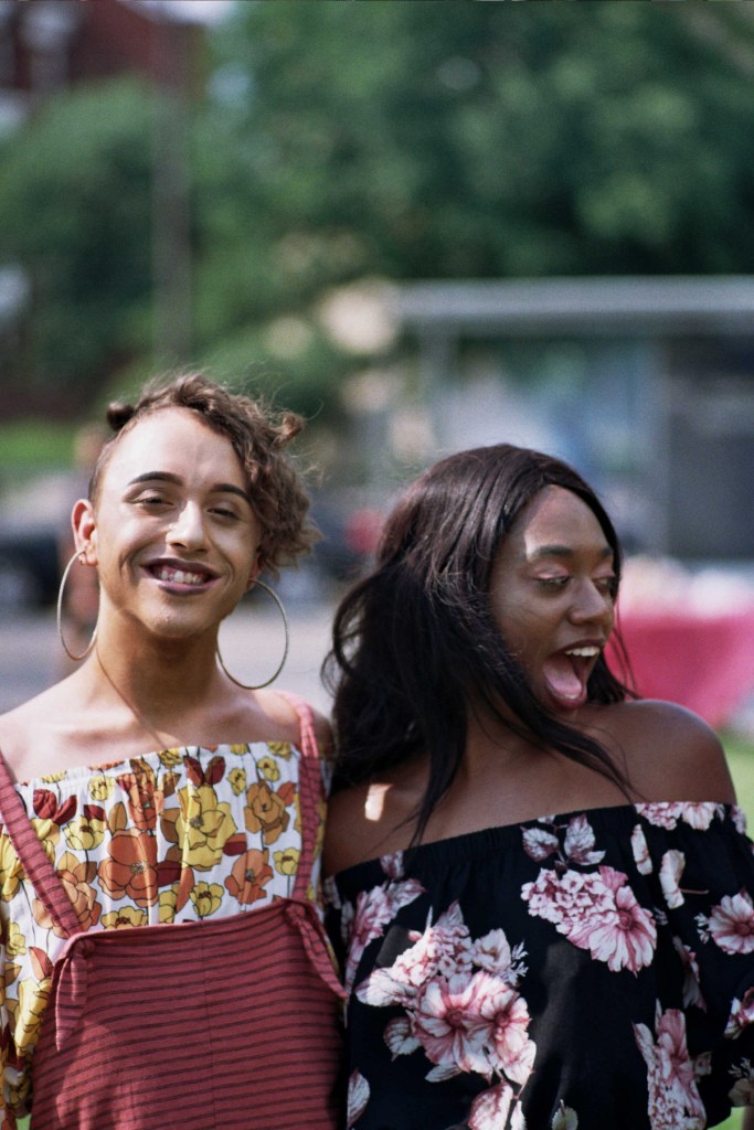 Two people walk arm-in-arm, grinning, wearing flowery dresses.