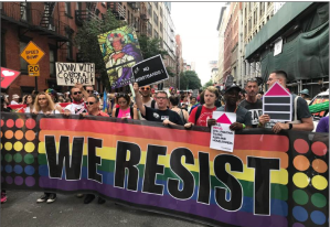 A large New York City protest march is led by a line of activists carrying a large rainbow-colored banner that reads "We Resist."